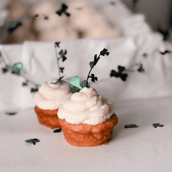 Zwei selbst gebackene Zitronen-Cupcakes mit hellem Topping und Kleeblatt-Dekoration auf einem weißen Tisch.